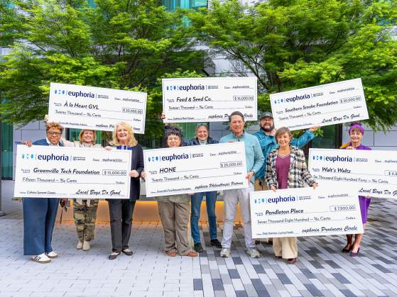 A group of people stand in front of a modern building, holding large checks from "euphoria" for various local organizations.