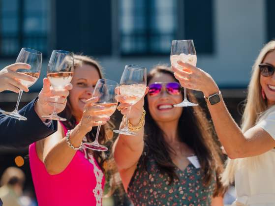 Four smiling people in the foreground raise glasses of rosé wine in a toast, with a blurred building and outdoor setting in the background.