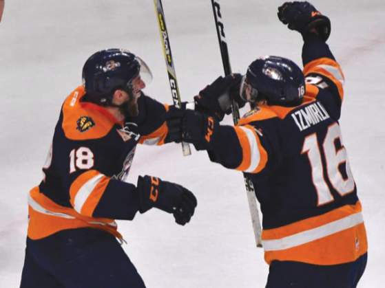 Two hockey players in orange jerseys celebrate a goal in the foreground, while a goalie in green and white and other players look on in the background.