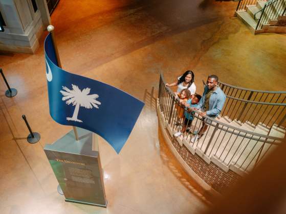Family of four on a staircase in the Upcountry History Museum gazing up at a sculpture of the South Carolina state flag.