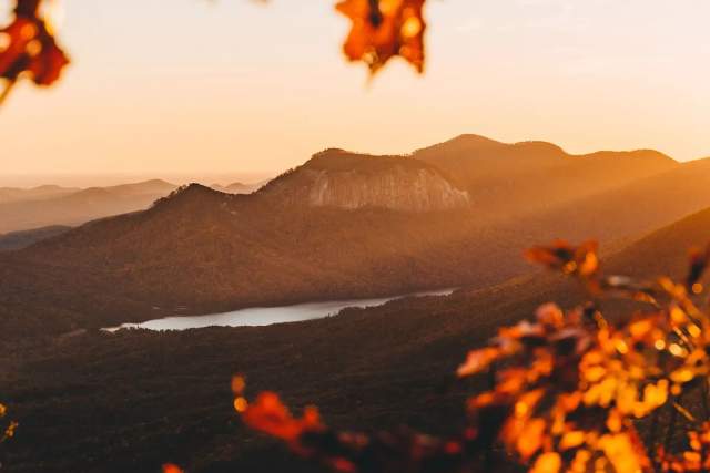 Autumn leaves in the foreground frame a sun-drenched mountain landscape with a lake in the distance.