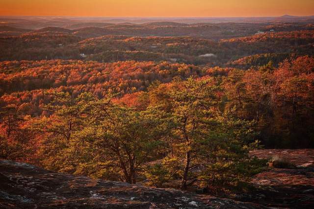 A rocky foreground overlooks rolling hills covered in autumn foliage under a warm sunset sky.