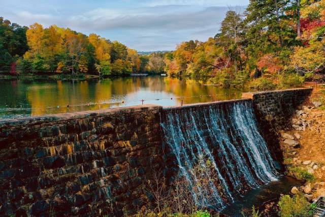 A stone dam with water cascading down its face separates a calm lake from a wooded landscape ablaze with autumn colors.