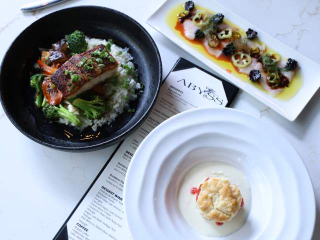 A black bowl of fish with rice and vegetables sits next to a white plate of thinly sliced fish and a white bowl with a biscuit in the foreground, all on a marble surface with a menu in the background.