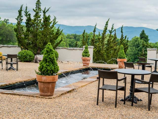 Exterior Italian-inspired gravel courtyard with black bistro tables, a rectangular long fountain, surrounded by several potted hedges overlooking the mountains on a sunny but slightly overcast day.