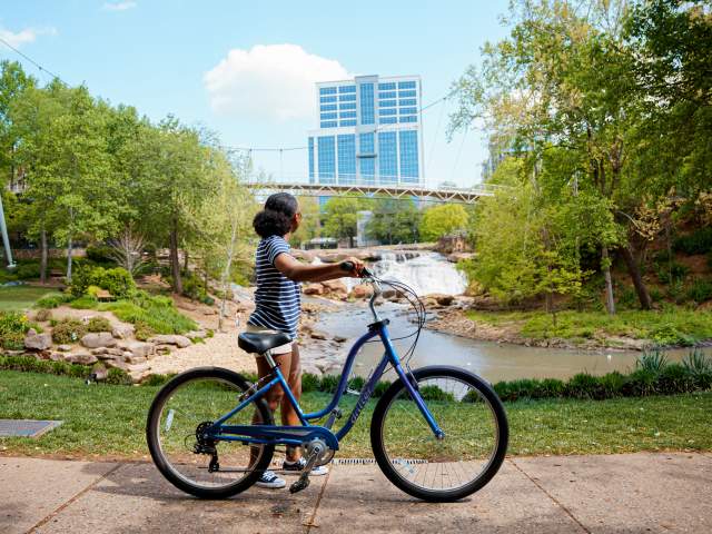 Woman with medium skin tone, blue and white striped shirt, and white shorts standing with a bike admiring the Liberty Bridge at Falls Park on the Reedy in Greenville, SC on a sunny summer day.