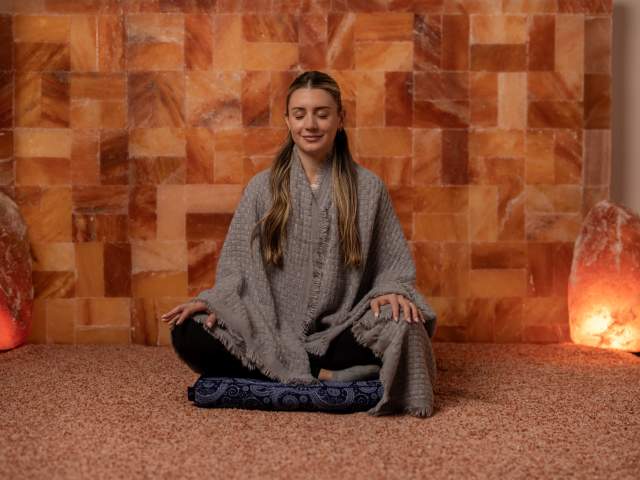 Woman sitting with legs crossed in a salt room at Raven & Moon with ambient warm lighting.