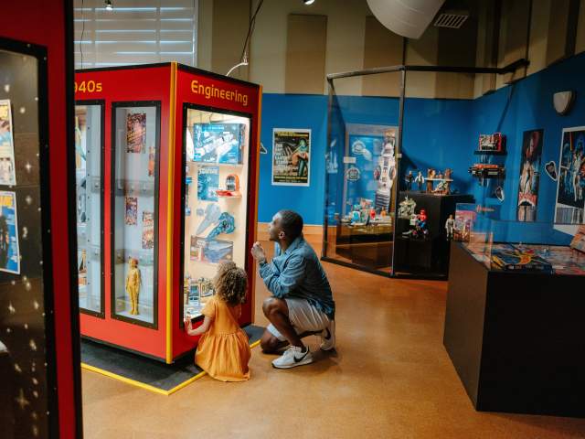 Man and little girl kneeling next to a lit up exhibit behind a glass enclosure that showcases vintage memorabilia from early movies or tv shows at the Upcountry History Museum.