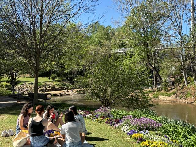 A group of young women having a picnic on a sunny spring day with flowers surrounding them in front of the Reedy River at Falls Park on the Reedy