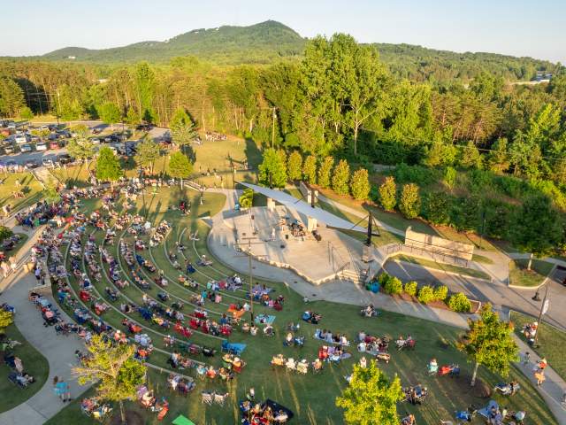 A crowd of people sits in a curved outdoor amphitheater in the foreground, with a lush forest and rolling green mountains in the background.