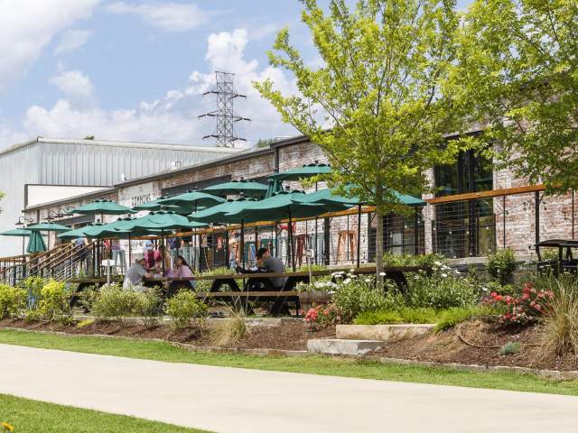 Yellow wildflowers line a paved path where cyclists ride past an outdoor patio with green umbrellas and a white building under a cloudy sky.