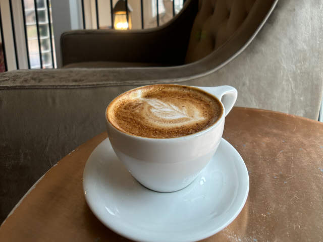 A latte with latte art sits on a copper table in front of a plush armchair, with a balcony and ornate ceiling visible in the background.