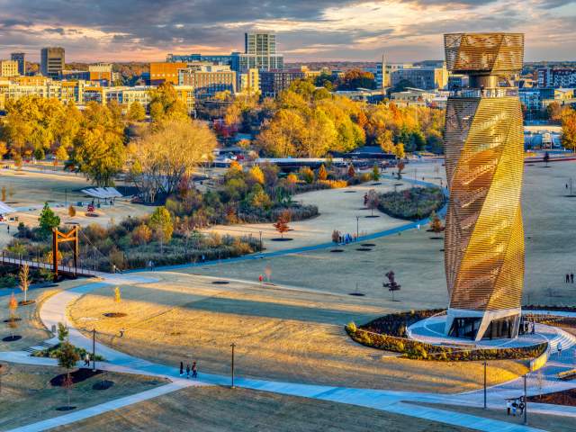 A tall, spiraling wooden tower stands in a grassy park foreground, with a city skyline and autumn trees in the background under a sunset sky.