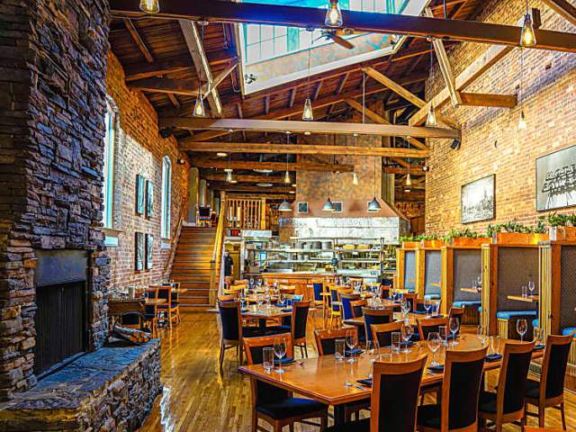 A rustic restaurant with a stone fireplace in the foreground and a bustling open kitchen in the background.