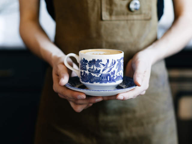 Hands in an apron hold a blue and white patterned cup of coffee on a matching saucer against a blurred dark background.