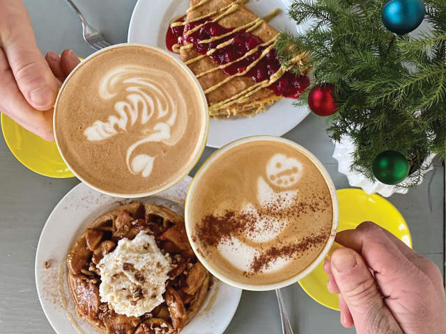 Two hands hold cups of latte art, one with a swirl and the other with a snowman, next to a waffle and a crepe, with a decorated Christmas tree in the background.