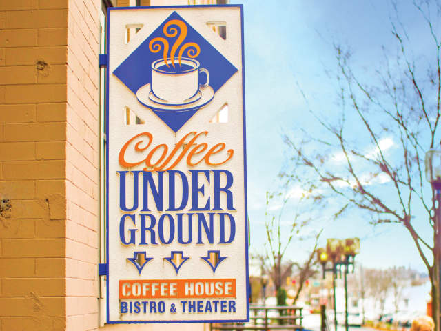 A sign for "Coffee Underground Coffee House Bistro & Theater" hangs on a brick building in the foreground, with a street scene and a cloudy sky in the background.