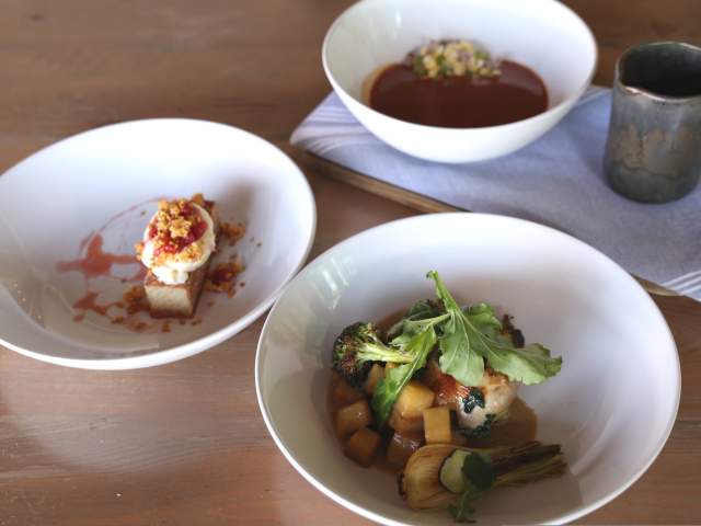 Three white bowls of food, one with dessert, one with a stew, and one with a main course, are arranged on a wooden table with a metal cup in the background.