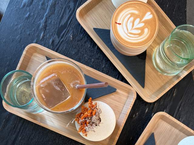 Two wooden trays with drinks and a glass of water sit on a dark wooden table, with one tray holding a latte with latte art and the other holding an iced coffee with a straw and a small decorative garnish.