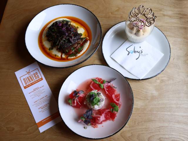 Three plates of food, including a dessert, are arranged on a wooden table next to a menu.