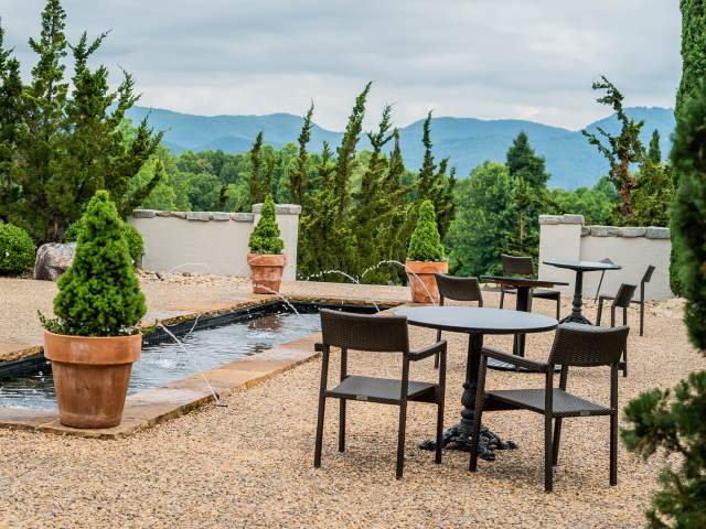 Outdoor seating with tables and chairs is arranged on a gravel patio next to a water feature, with lush green trees and rolling mountains in the background.