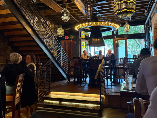 In the foreground, people are seated at tables and a staircase leads up to a mezzanine, while the background reveals a bar area with patrons and decorative lighting.