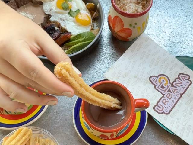 A hand dips a churro into a mug of hot chocolate in the foreground, with a plate of food and other churros in a cup visible on a table in the background.