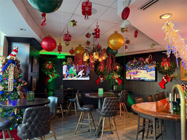 A festive Christmas-decorated restaurant with a decorated tree in the foreground and holiday ornaments and screens displaying holiday scenes in the background.