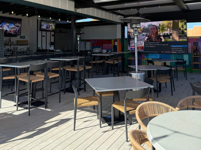 In the foreground, empty tables and chairs are set up on a wooden deck, while the background features a bar area and a large outdoor screen displaying a video. (The Slice - Simpsonville)