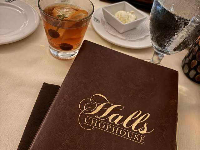 A Halls Chophouse menu resting on a crisp white tablecloth, accompanied by a crafted cocktail with vibrant garnishes, capturing the fine dining ambiance.
