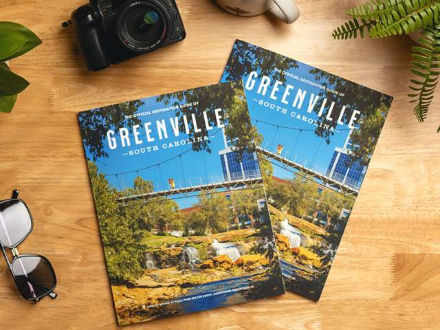 Two copies of the 2025 Official Destination Guide of Greenville, SC, sit on a counter next to a camera, sunglasses, coffee, and a plant.