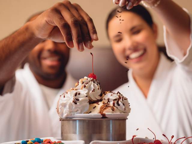 Man and woman sprinkling toppings onto a giant ice cream sundae.