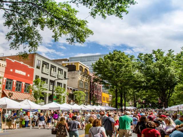 Downtown Greenville farmers market with many vendors set up and a large crowd of shoppers walking by the stands.