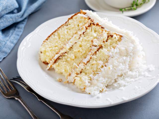 A slice of coconut cake on a white plate atop a blue tablecloth.