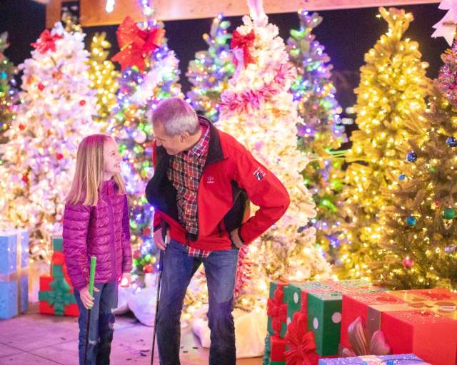 A dad and daughter stand amongst well-lit Christmas trees at Takeoff Mini Golf in Greenville, SC.