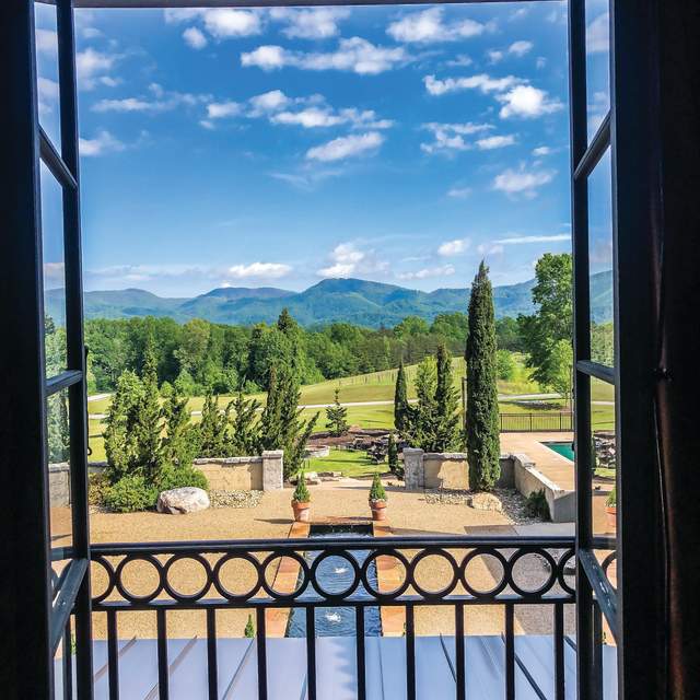 View of mountains in the distance with some scattered clouds and trees on a sunny day from looking out a hotel room door with a small balcony that has minimally decorative iron railing.