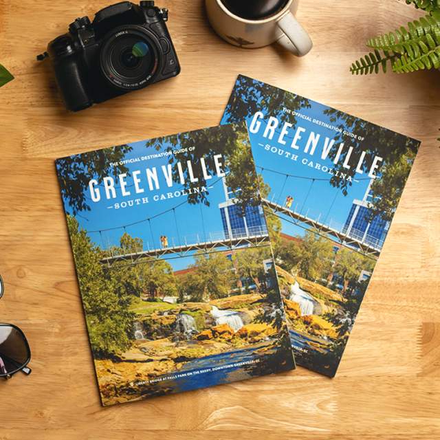 Two copies of the 2025 Official Destination Guide of Greenville, SC, sit on a counter next to a camera, sunglasses, coffee, and a plant.