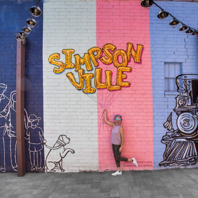 A pink, white, and purple mural in Simpsonville, SC that lets a person interact with it by posing like you're holding balloons.