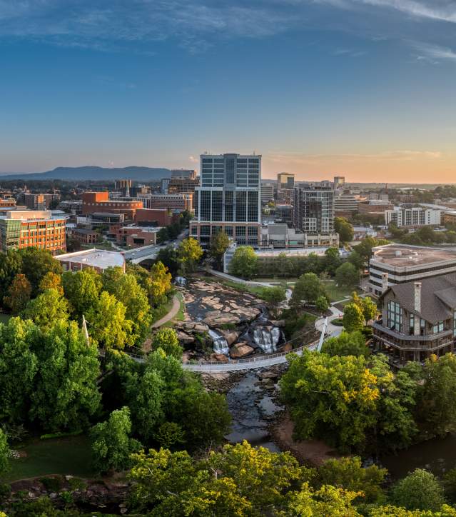 A lush green park with a flowing river and waterfalls in the foreground leads to a city skyline under a bright sunset in the background.