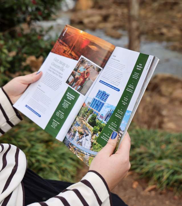 A person in a black and white striped shirt reading the 2026 Destination Guide to Greenville, SC in Falls Park on the Reedy.