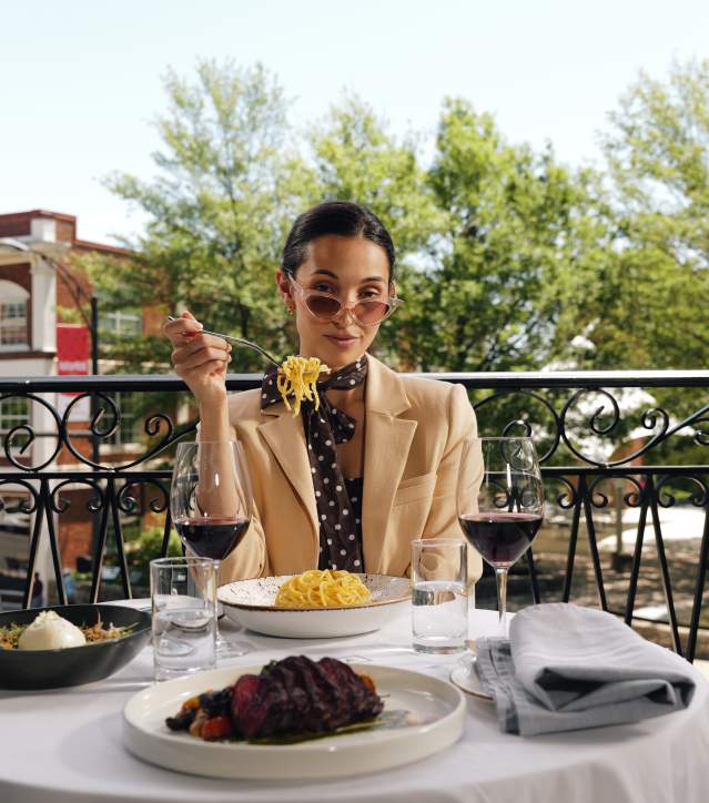 A woman in a beige blazer eats pasta at a white-clothed table on a balcony, with green trees and a brick building in the background.