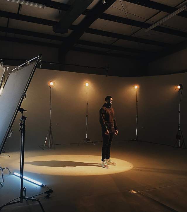 Male subject with yellow tinted, soft spotlight on him casting a shadow in a dark studio with several other yellow, soft lights behind him and film equipment.