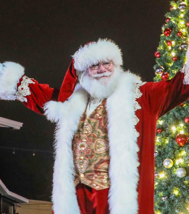 Santa Claus with arms outstretched stands in the foreground before a brightly lit Christmas tree and a crowd of onlookers in the background.