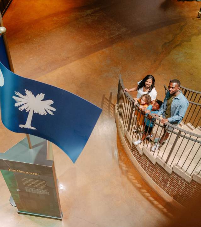 Family of four on a staircase in the Upcountry History Museum gazing up at a sculpture of the South Carolina state flag.