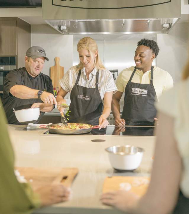 Group of men and women at a cooking class with a chef demonstrating how to prepare a dish on a stove.