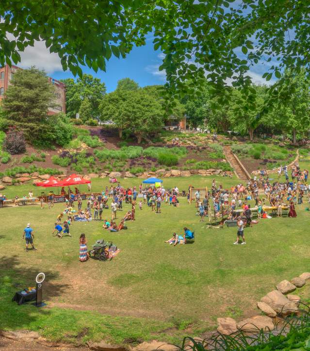A bustling crowd in Falls Park during Artisphere, gathered near a stage for live performances, with the park's natural beauty providing a scenic backdrop to the festival.