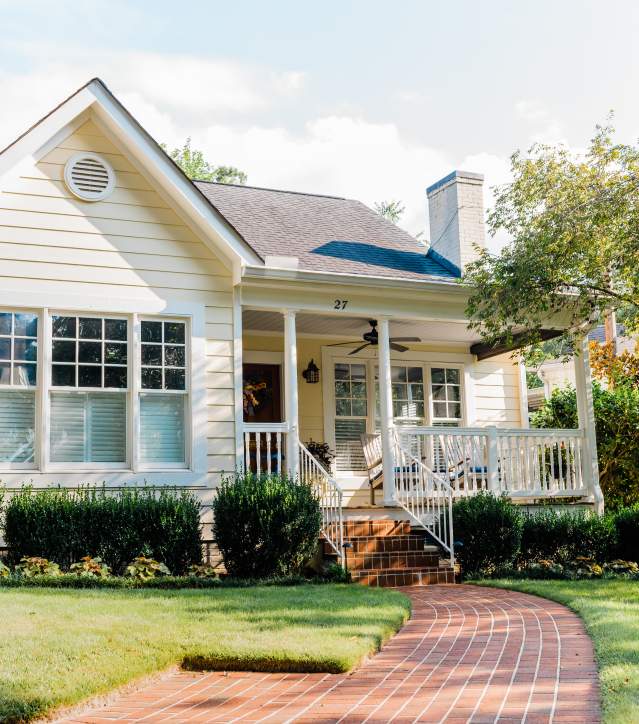A cozy yellow house with a brick pathway, framed by a well-kept lawn and luscious hedges.