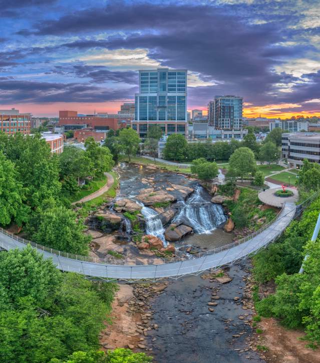 An aerial view of downtown Greenville, featuring the iconic Liberty Bridge spanning over Falls Park, with lush greenery and urban buildings in the background.