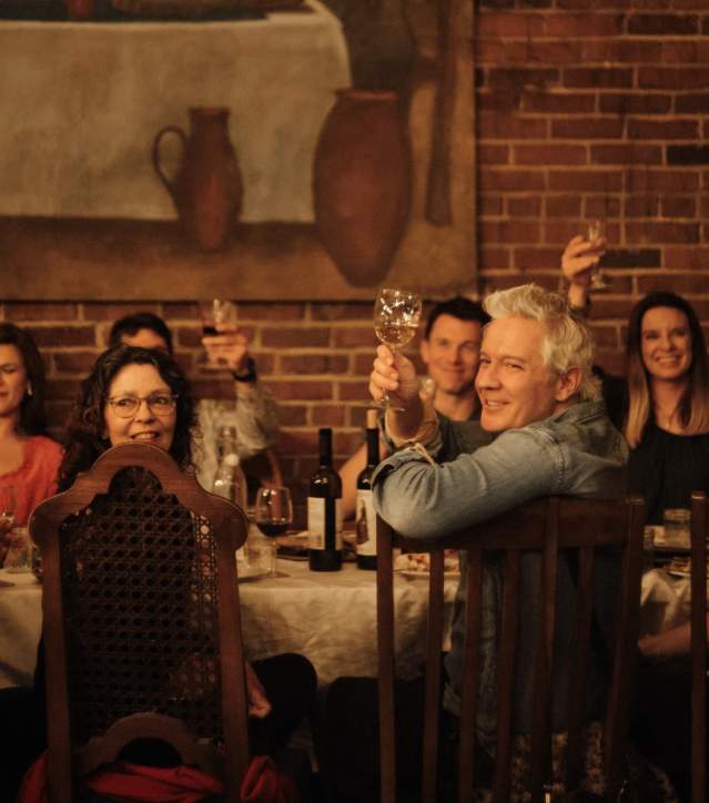 A lively group of people raising their glasses in a toast at Keipi, enjoying a toasting dinner inspired by Georgian traditions, with a table full of delicious food and wine.