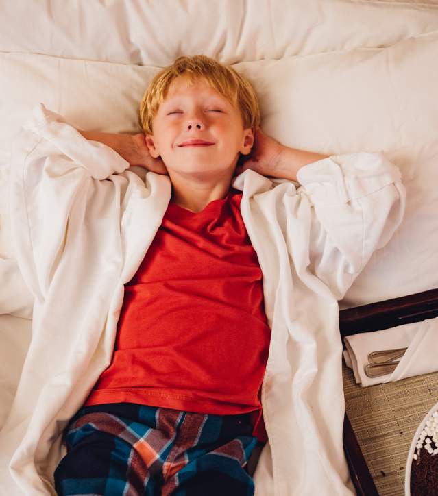 Boy laying on fluffy hotel bed with pizza and ice cream next to him.